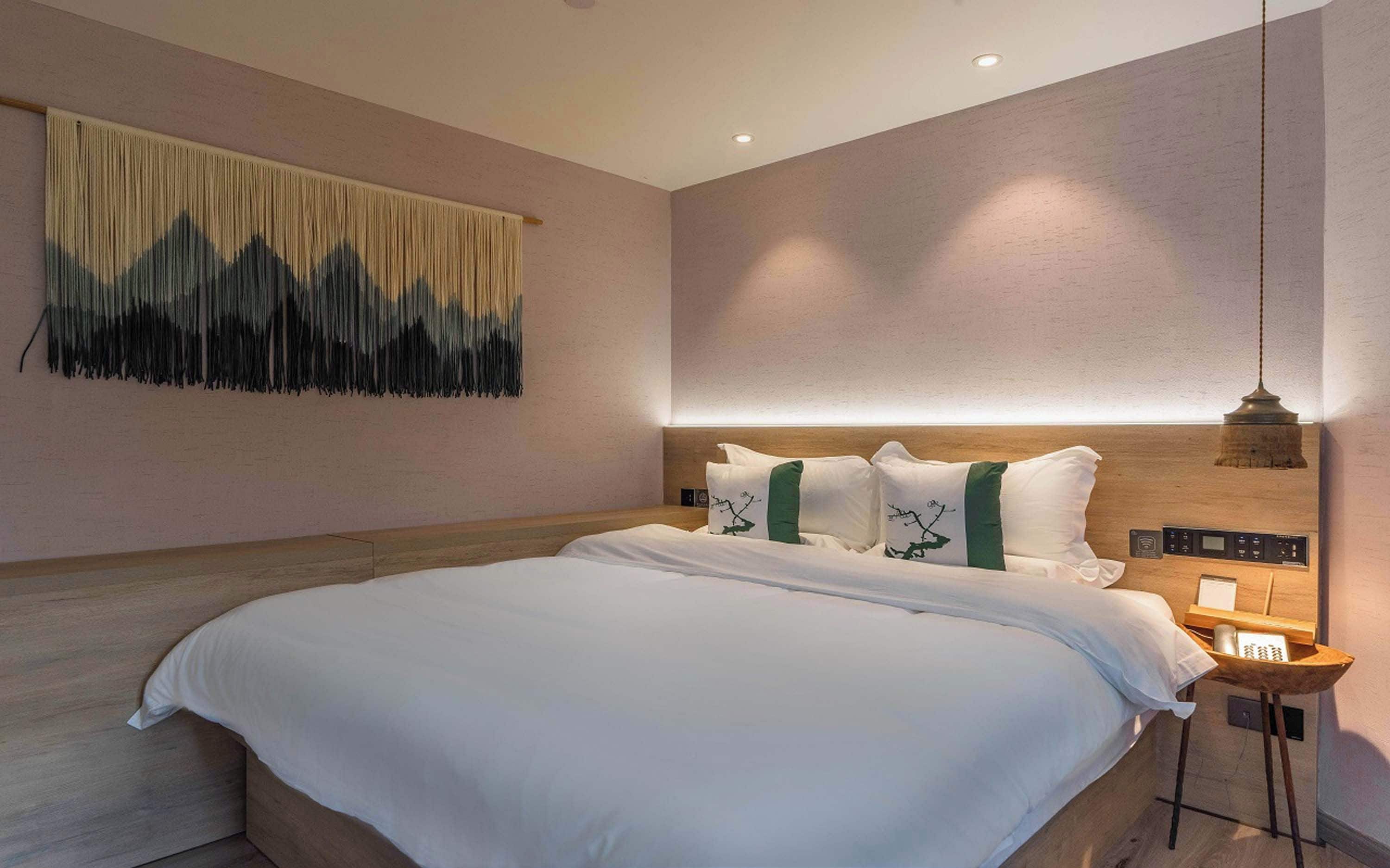 Una habitación de hotel moderna con una cama grande, un cabecero con controles integrados, una mesita de noche con lámpara y una obra de arte colgada en la pared.