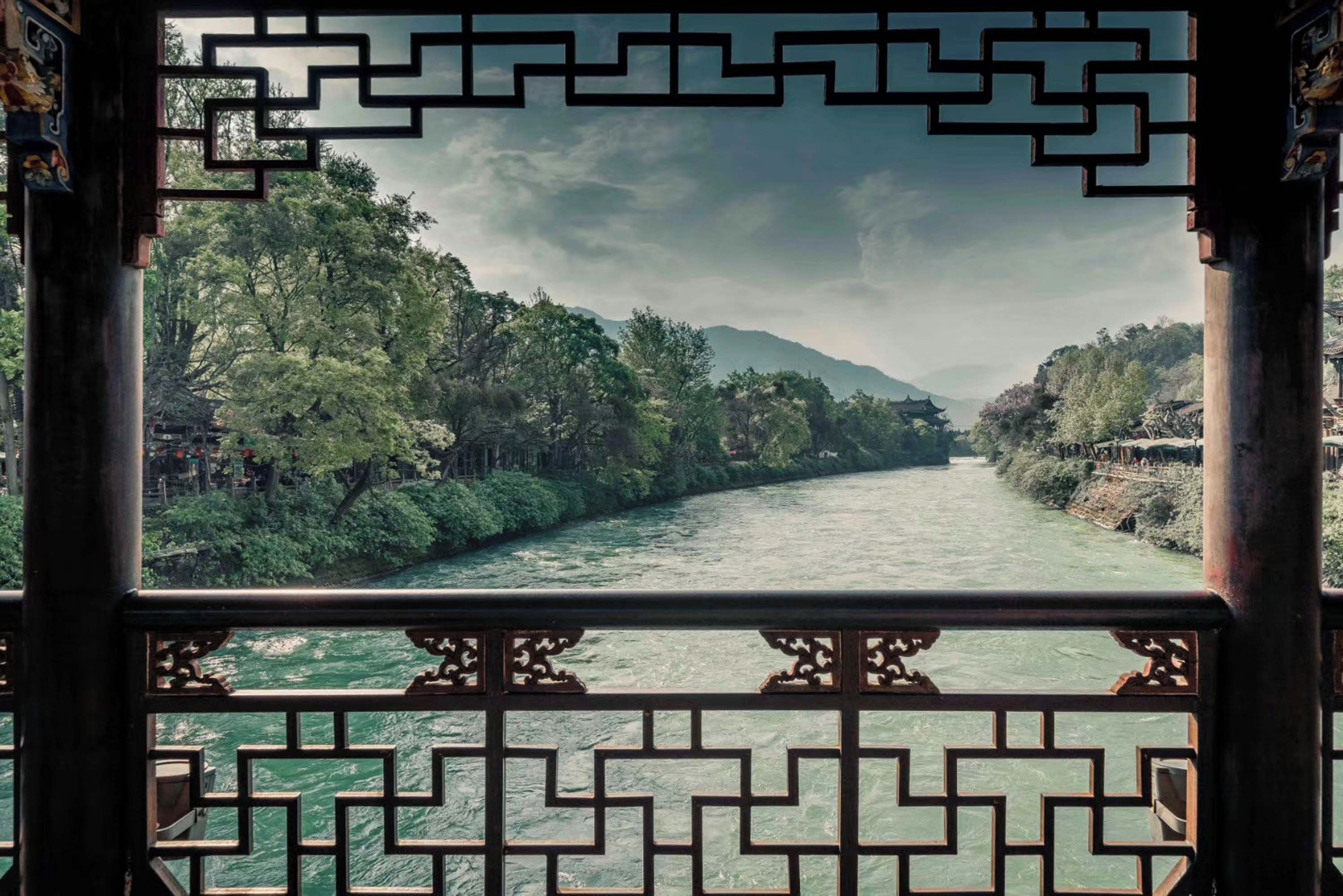 A view through a traditional Chinese window with geometric patterns, overlooking a river flanked by lush greenery and distant hills.