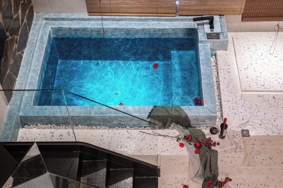 Piscina com borda de pedra, uma rosa vermelha e uma garrafa de vinho ao lado.