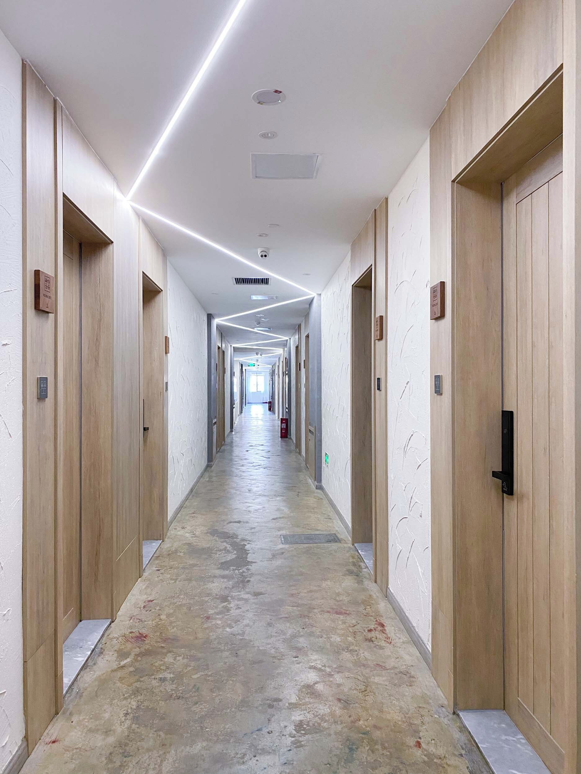 Un largo pasillo de hotel, estrecho, con puertas de madera a ambos lados, suelo con diseño y luces empotradas.