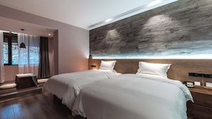 Ein modernes Hotelzimmer mit zwei Betten, einem großen Fenster mit Vorhängen, Holzboden und einer an der Wand montierten Bedieneinheit.
