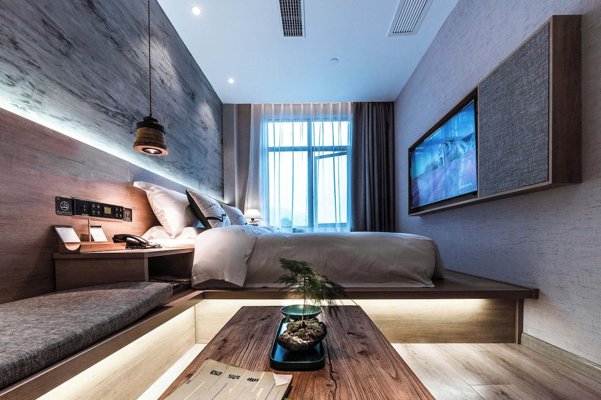Quarto de hotel moderno com cama, mesa de cabeceira, abajur e uma janela grande.