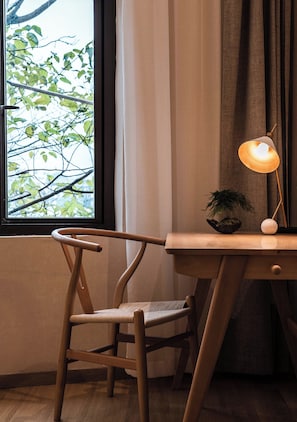 Uma cadeira de madeira com assento de fibra natural, uma mesa com luminária e vista para árvores pela janela.