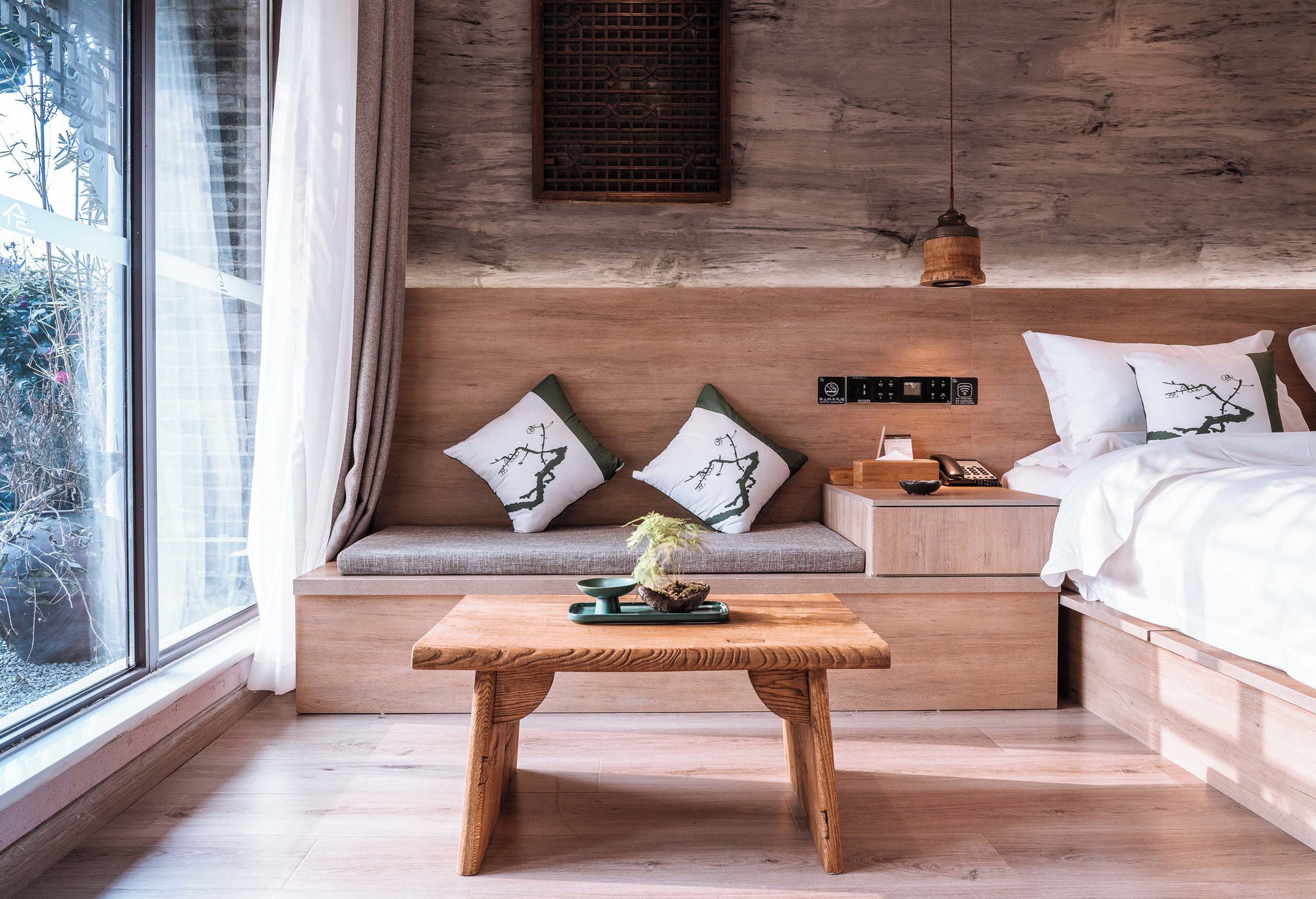 A modern bedroom with a wooden headboard, a bedside table with a lamp, and a view of greenery outside.