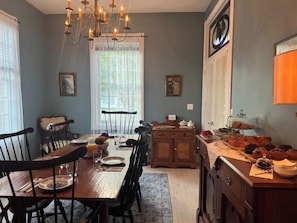 Een eetkamer met een houten tafel voor vier personen, omringd door stoelen, een kroonluchter en een raam met dunne gordijnen.