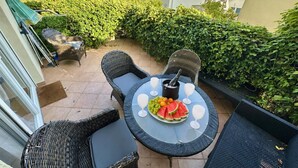 Une terrasse extérieure avec des meubles en osier, une table garnie de fruits et une bouteille, et une vue sur une colline.