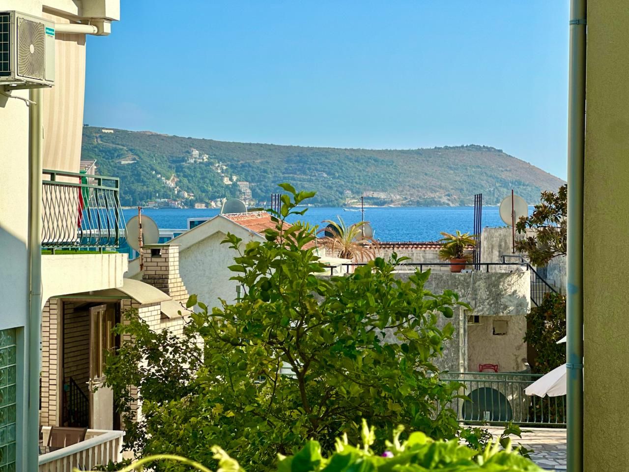 A coastal residential area with buildings, a view of the sea, and a hill in the background.