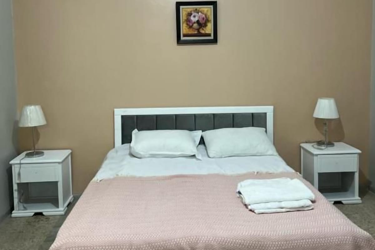 A bedroom with a bed, two bedside tables, a nightstand, a wall-mounted picture, and a carpeted floor.