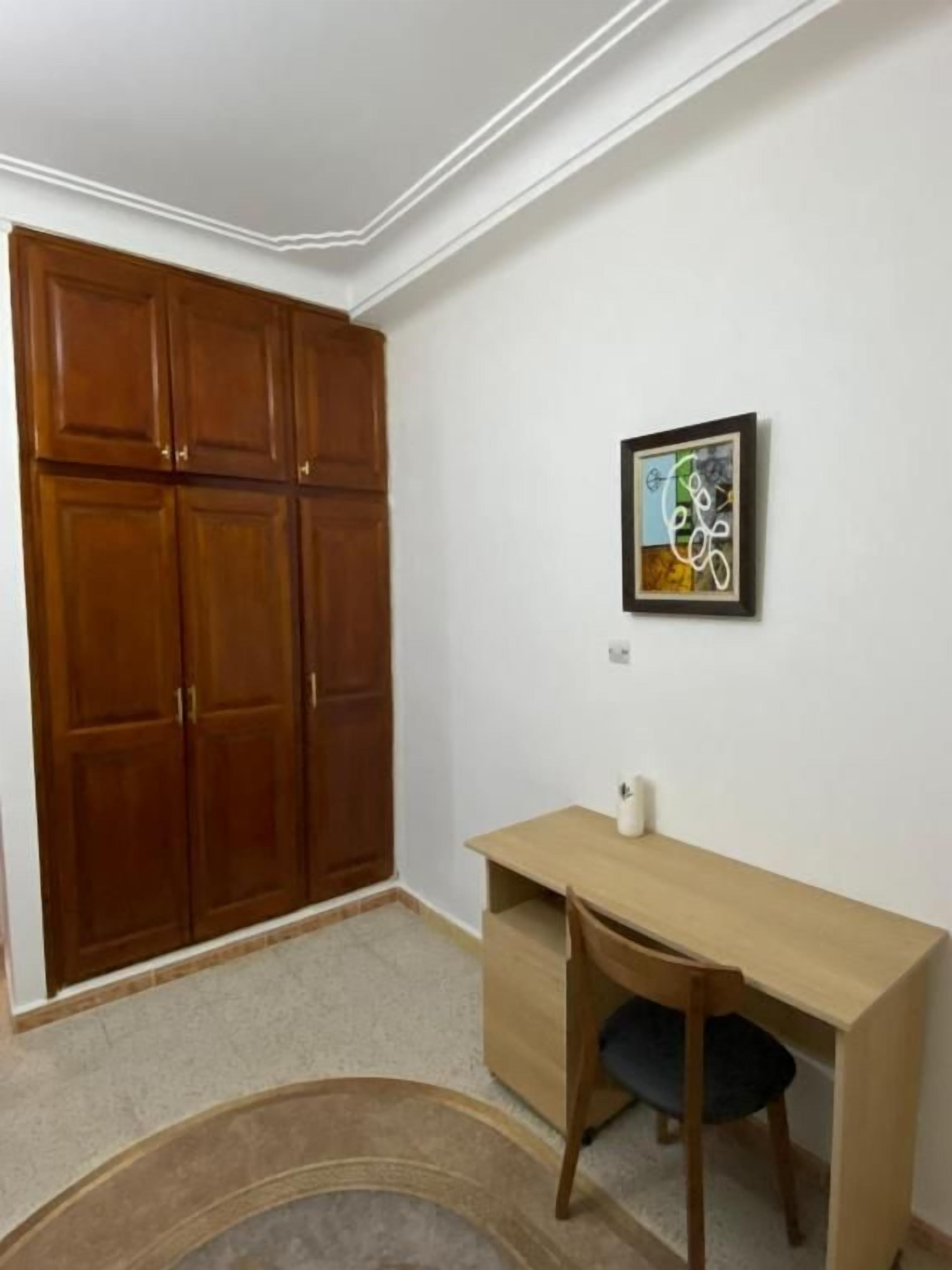 A room with a wooden wardrobe, a desk with a chair, and a framed picture on the wall.