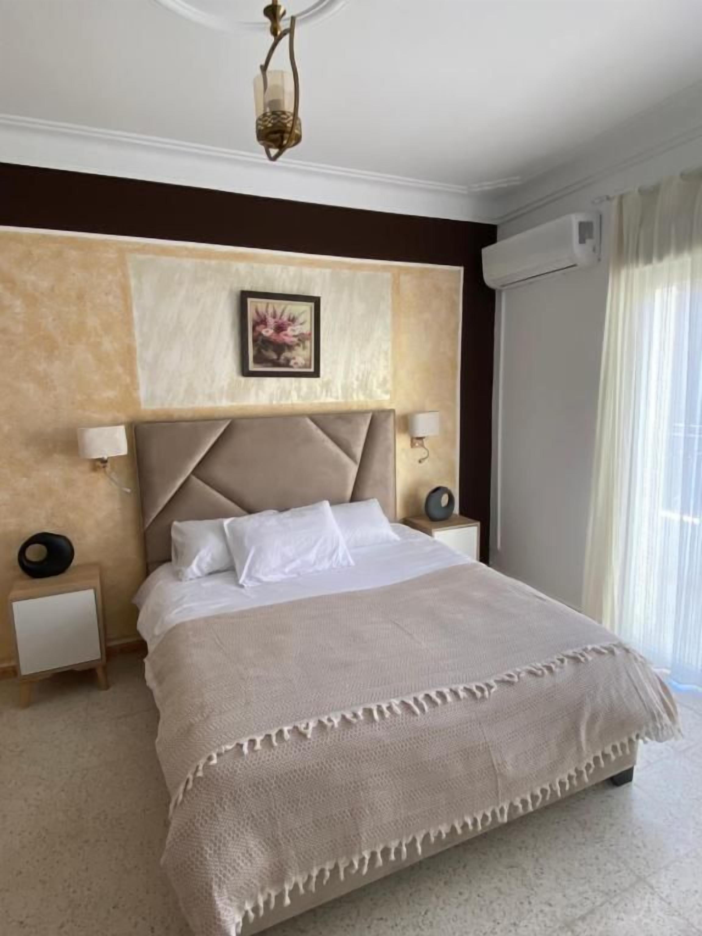 Habitación de hotel con una cama grande, cabecera, dos mesitas de noche, aire acondicionado de pared y una ventana con cortinas.