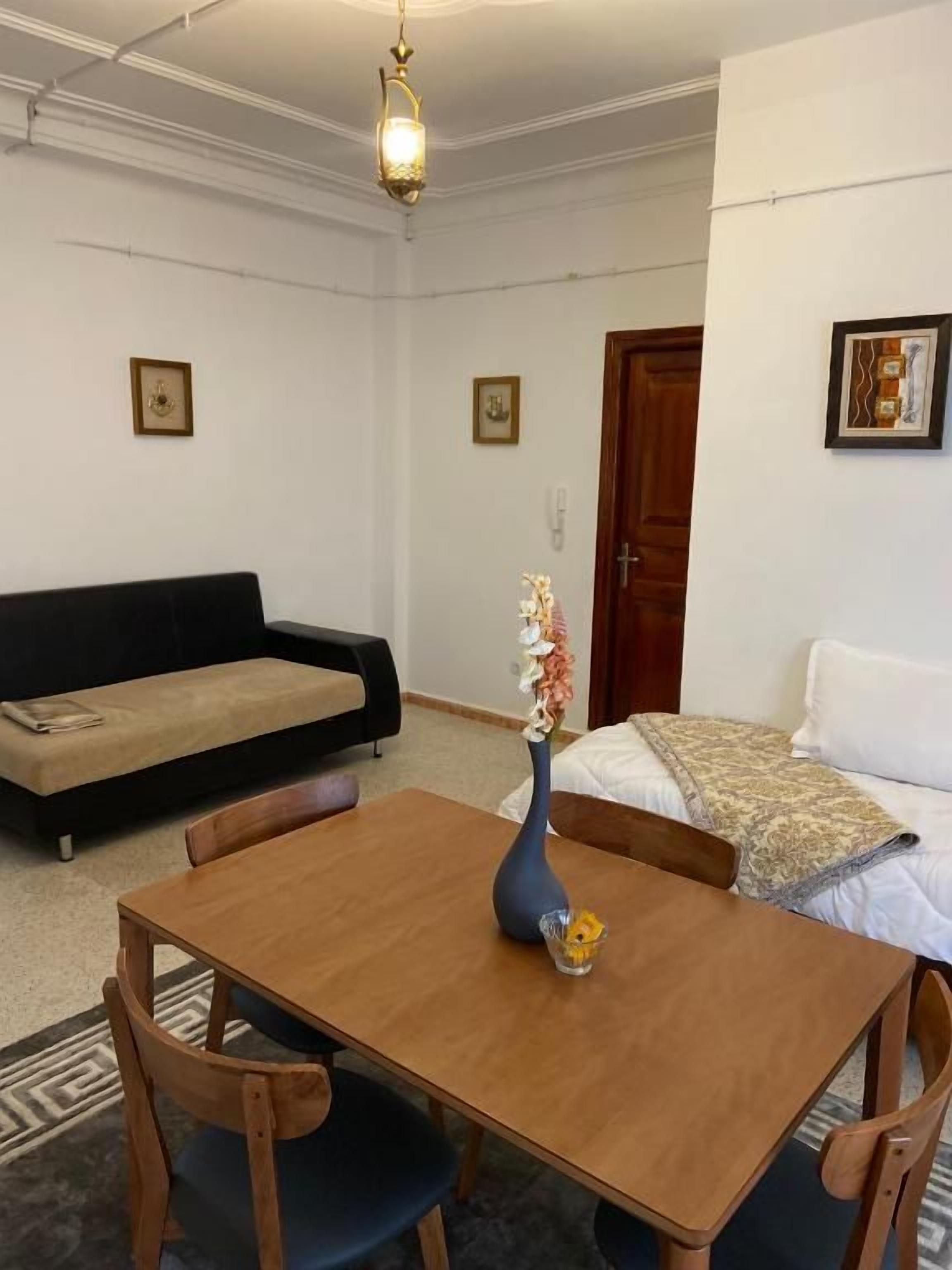 Una habitación con una mesa de comedor de madera, dos sillas, un sofá, una cama y un florero con flores.