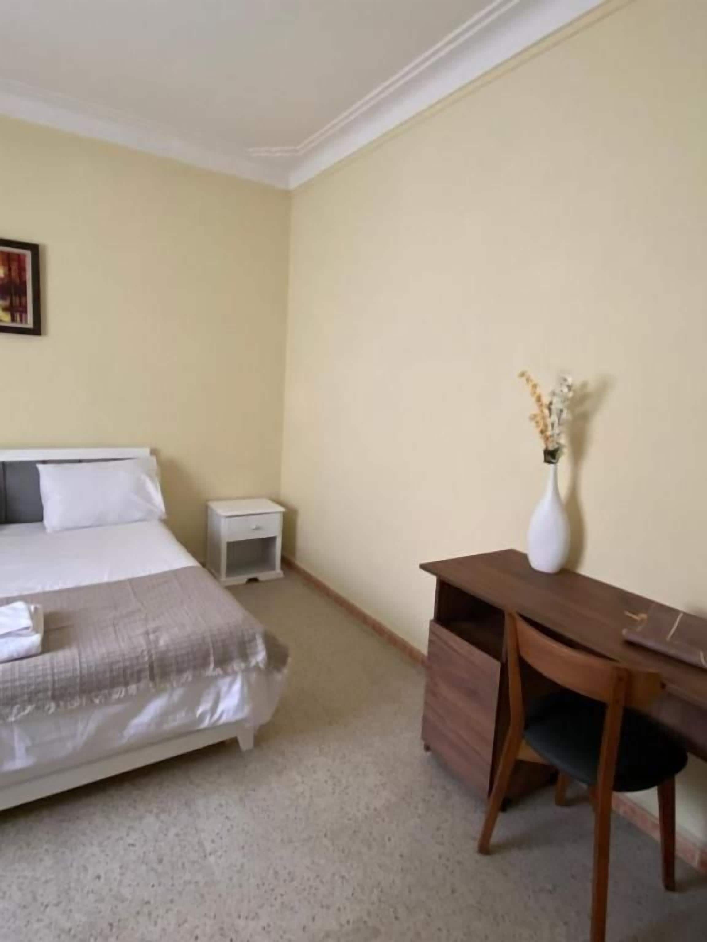A hotel room with a bed, bedside table, desk, chair, and a vase with flowers.