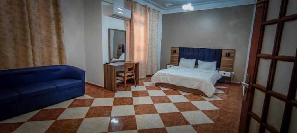 A hotel room with a checkered floor, a bed with white bedding, a blue sofa, a desk with a chair, and a mirror.