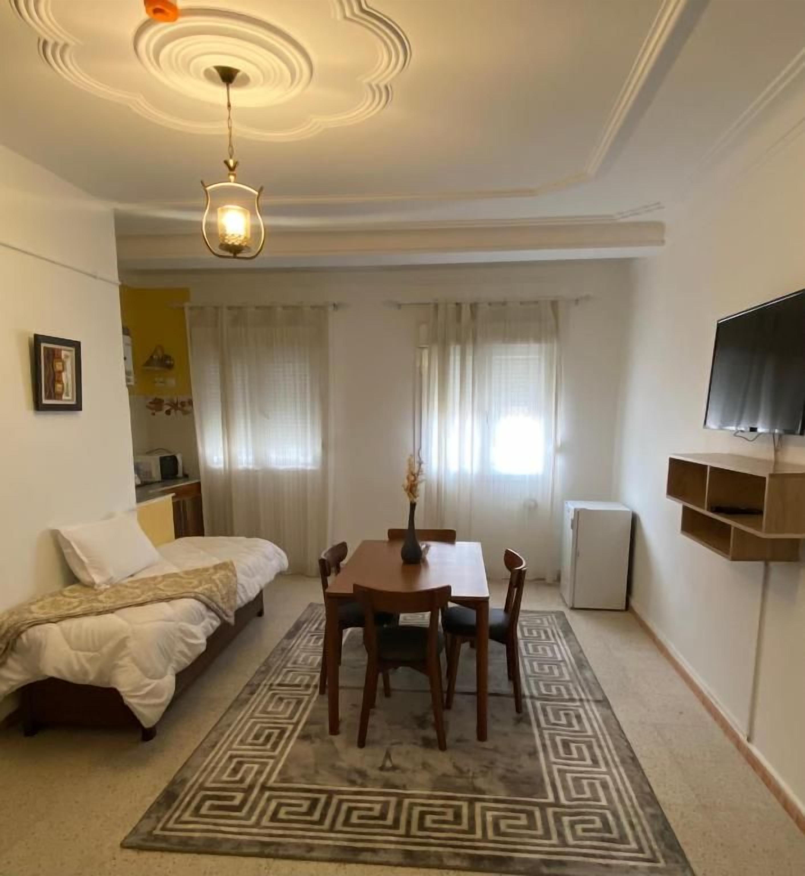 Habitación compacta con cama, una mesa de comedor con sillas, un televisor pequeño y un tapete decorativo.