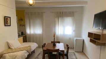 Habitación compacta con cama, una mesa de comedor con sillas, un televisor pequeño y un tapete decorativo.