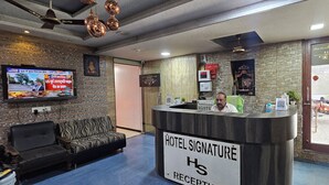 Zona de recepción de un hotel con mostrador, televisor mostrando noticias, sofá y pared con diseño de piedra.