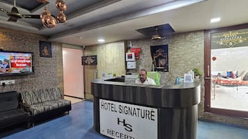 Un espace d’accueil d’hôtel comprenant un comptoir de réception, un téléviseur diffusant les actualités, un canapé et un mur orné d’un motif en pierre.