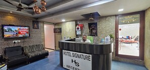 Zona de recepción de un hotel con mostrador, televisor mostrando noticias, sofá y pared con diseño de piedra.