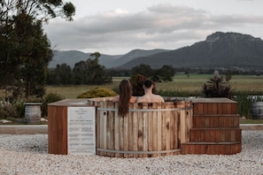 Two people in a wooden hot tub with a sign listing safety rules, surrounded by a gravel area and a view of a mountain. - The Wellness Farm Mindful Villa 5 (Broke)