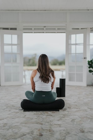 A person seated on a yoga mat in a room with large windows and doors, a speaker, and a plant. - The Wellness Farm Mindful Villa 5 (Broke)
