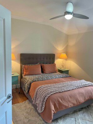 A bedroom with a bed, two bedside lamps, a ceiling fan, and a door. - The Wellness Farm Hamptons Cottage (Broke)
