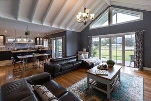 A spacious living area with a dark-colored sofa, a coffee table, and a dining table. There is a kitchen in the background with modern appliances and a large window. - The Wellness Farm Hamptons Cottage (Broke)