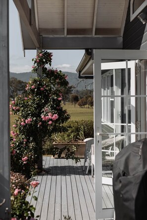 A wooden deck with a pergola, a white wicker chair, and a rose bush. - The Wellness Farm Hamptons Cottage (Broke)
