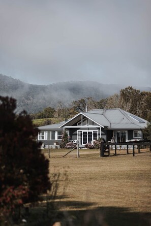 A two-story house with a large front yard, a covered porch, and a view of a hilly landscape. - The Wellness Farm Hamptons Cottage (Broke)