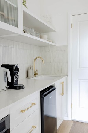 A modern kitchen with white cabinetry, a black dishwasher, a coffee maker, and a gold faucet. - The Wellness Farm Restore Villa 6 (Broke)