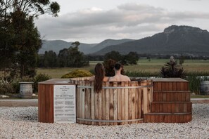 Two people in a wooden hot tub with a sign listing safety rules, surrounded by a gravel area and a view of a mountain. - The Wellness Farm Restore Villa 6 (Broke)
