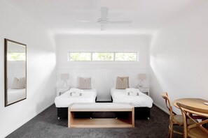 Habitación de hotel con dos camas, una mesita pequeña, una silla, un ventilador de techo y una ventana con vistas a zonas verdes.