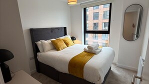 Ein modernes Schlafzimmer mit Bett, Nachttischlampe, Fenster mit Blick auf Gebäude und Spiegel an der Wand.