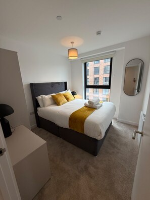Un dormitorio moderno con cama, lámpara de noche, ventana con vista a edificios y un espejo en la pared.