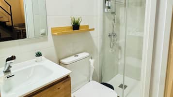 Un baño con un lavamanos blanco, tocador de madera, inodoro, mampara de ducha de vidrio y una pequeña planta enmacorada en una repisa.