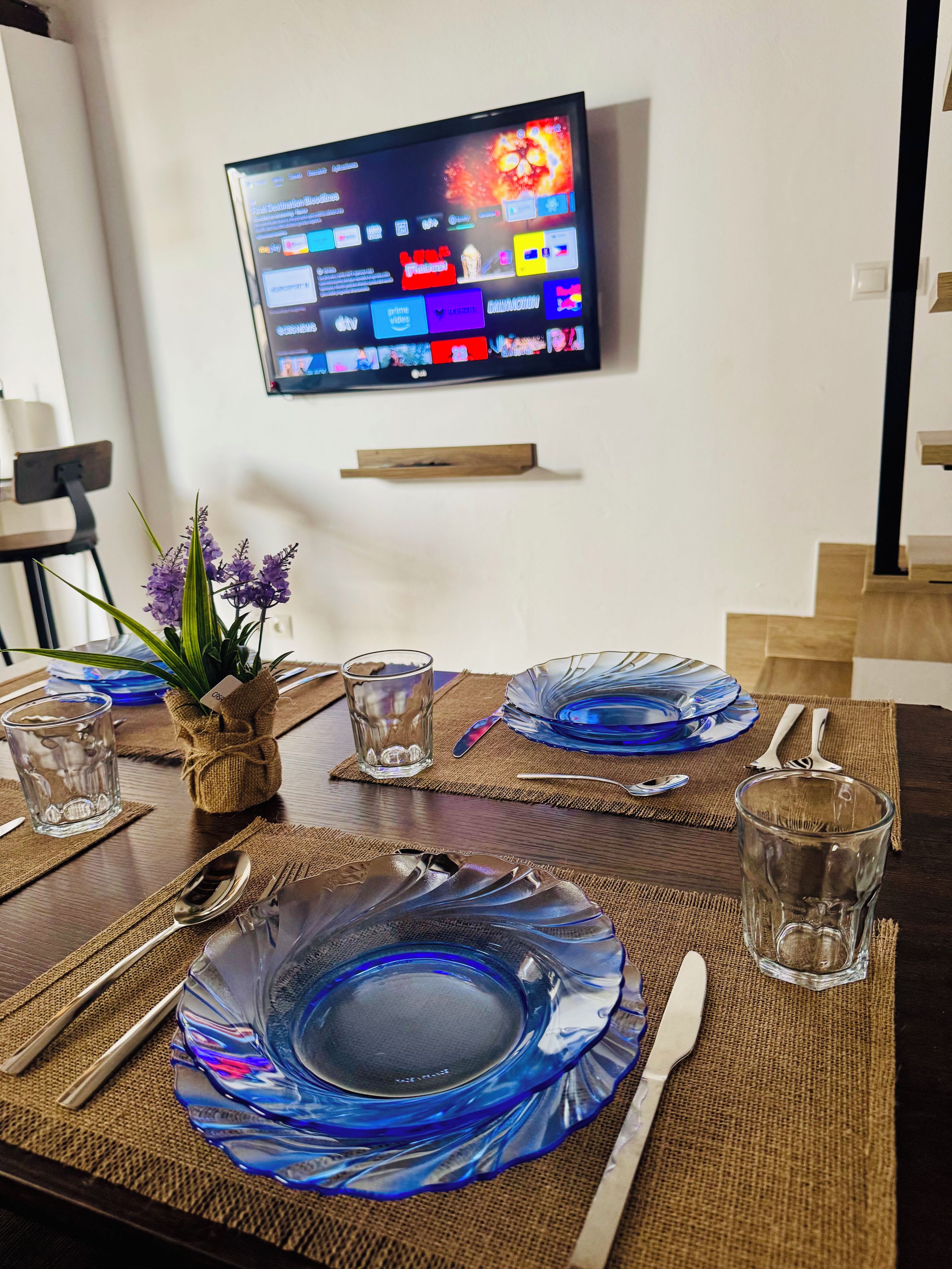 Une table à manger dressée avec de la vaisselle en verre bleue, un téléviseur fixé au mur et une petite plante posée sur la table.