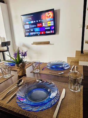 A dining table set with blue glassware, a TV mounted on the wall, and a small plant on the table. - "Your Charming Retreat" Apartment with a Mezzanine in Alegría de Jerez (Jerez de la Frontera)