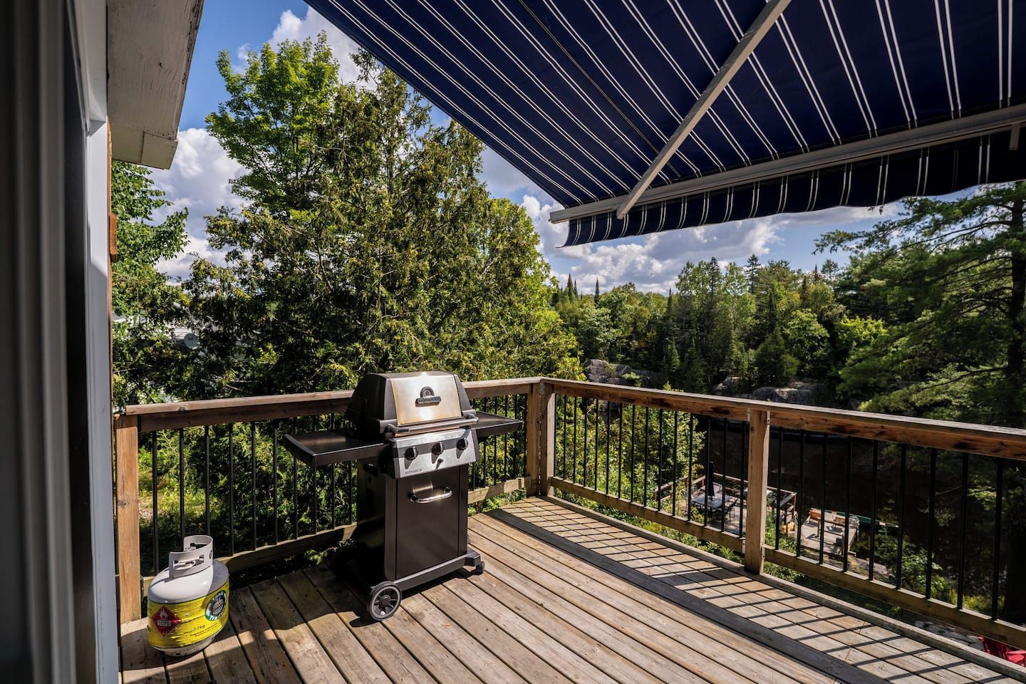 Eine Holzterrasse mit Grill und Propangasflasche, umgeben von einem Metallgeländer und Blick auf Bäume.