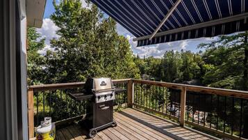 Eine Holzterrasse mit Grill und Propangasflasche, umgeben von einem Metallgeländer und Blick auf Bäume.