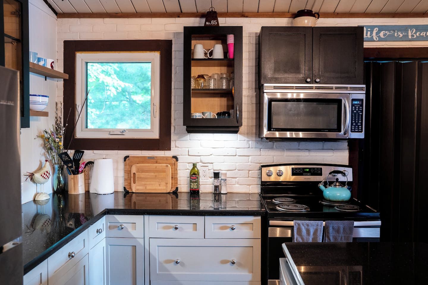 A kitchen with white cabinets, a black countertop, a window, a microwave, a stove, and a kettle.