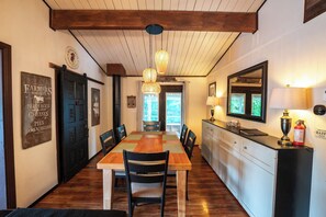 Un espace repas avec une table et des chaises en bois, une cuisine dotée d’un comptoir et de placards, et un plafond avec des poutres apparentes.