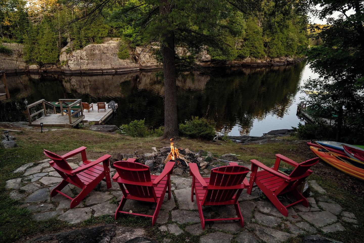 Un paisaje junto al lago con sillas Adirondack rojas, un muelle y kayaks.