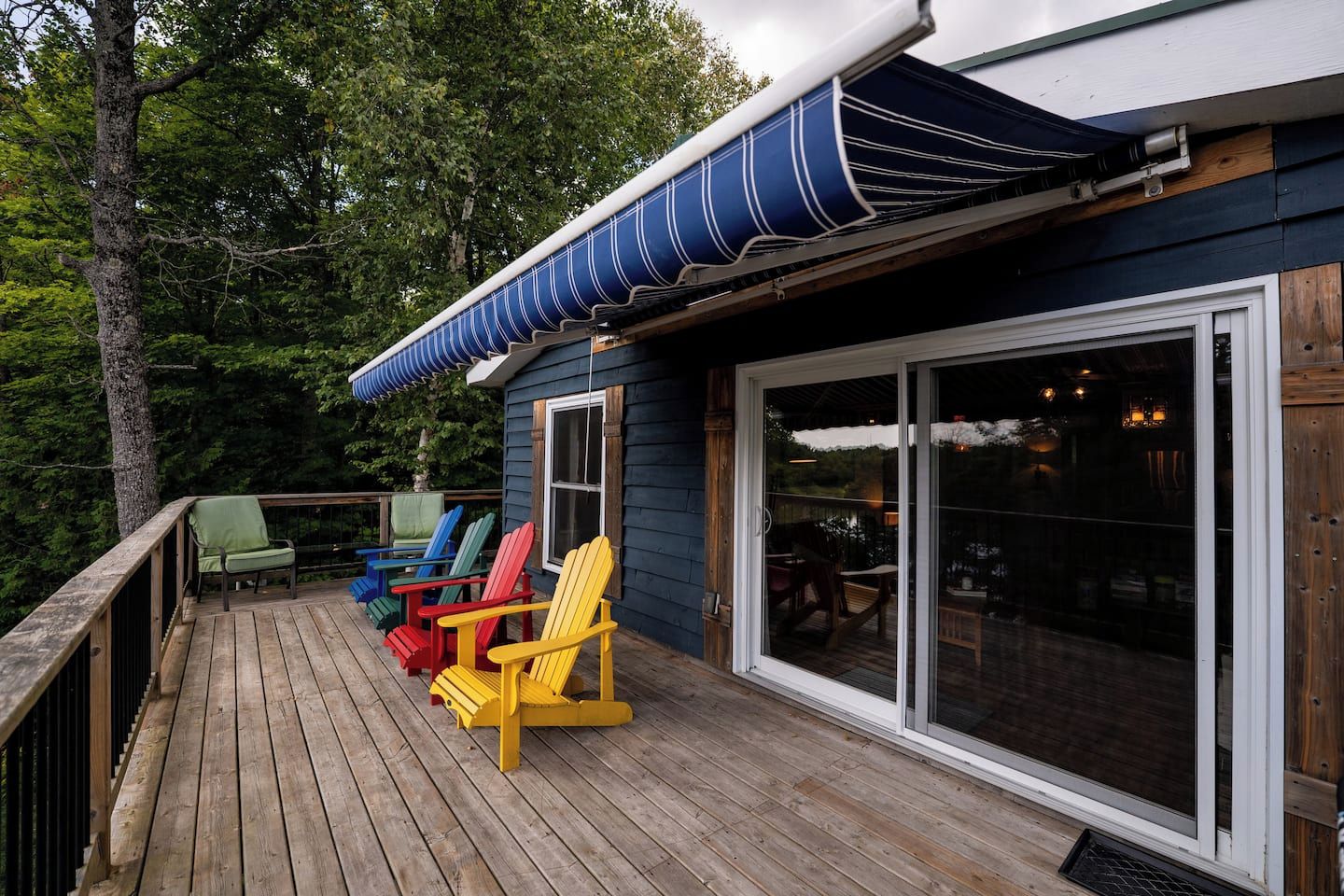 Una terraza de madera con sillas Adirondack de colores, un área cubierta con toldo a rayas y un amplio ventanal que da a un espacio interior.