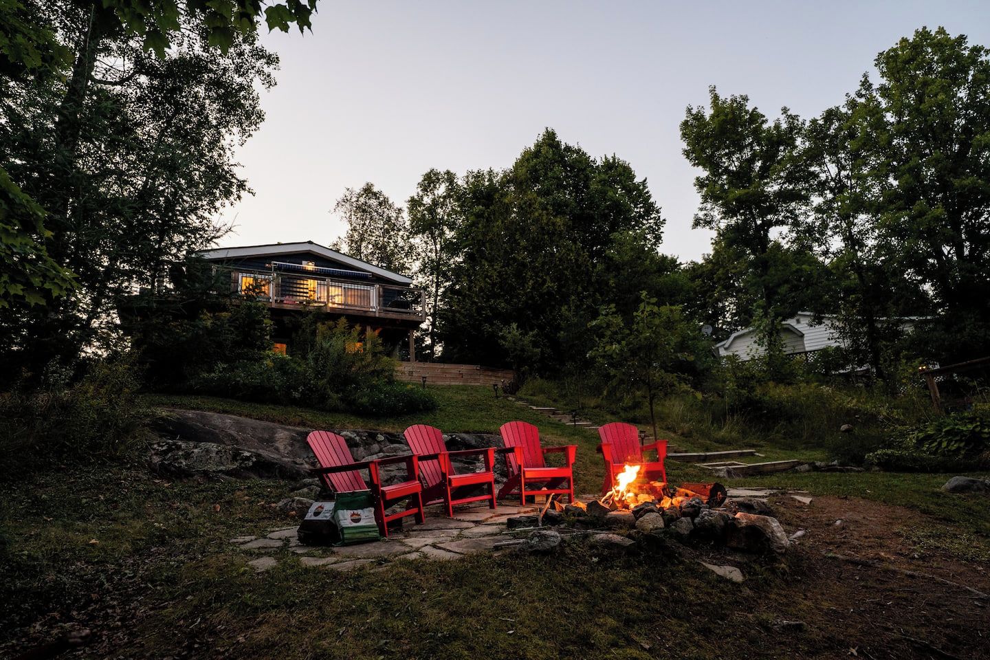 Eine Terrasse mit roten Adirondack-Stühlen und einer Feuerstelle, umgeben von einer natürlichen Landschaft und einem Haus im Hintergrund.