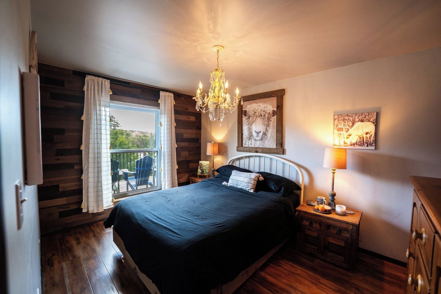 A bedroom with a large bed, a chandelier, a balcony with chairs, and a painting on the wall.