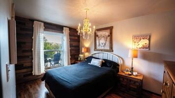 A bedroom with a large bed, a chandelier, a balcony with chairs, and a painting on the wall.