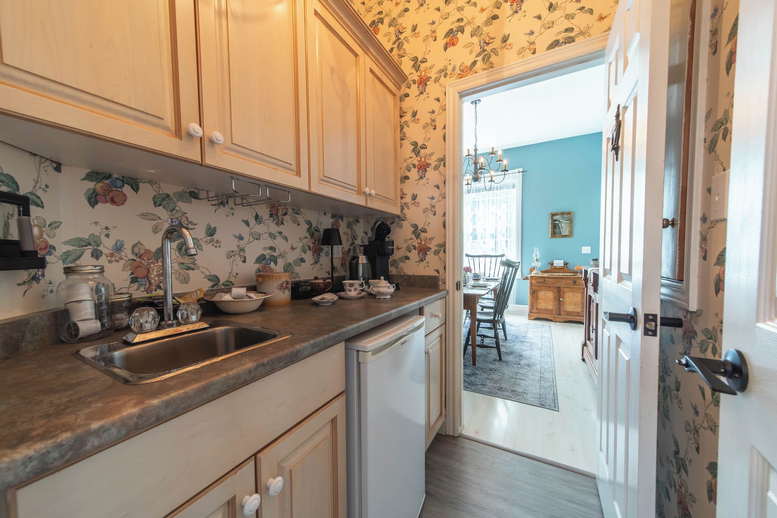 Una cucina con mobili in legno, un lavello e un piano di lavoro con diversi oggetti. Si apre una porta verso una zona pranzo con tavolo e sedie.