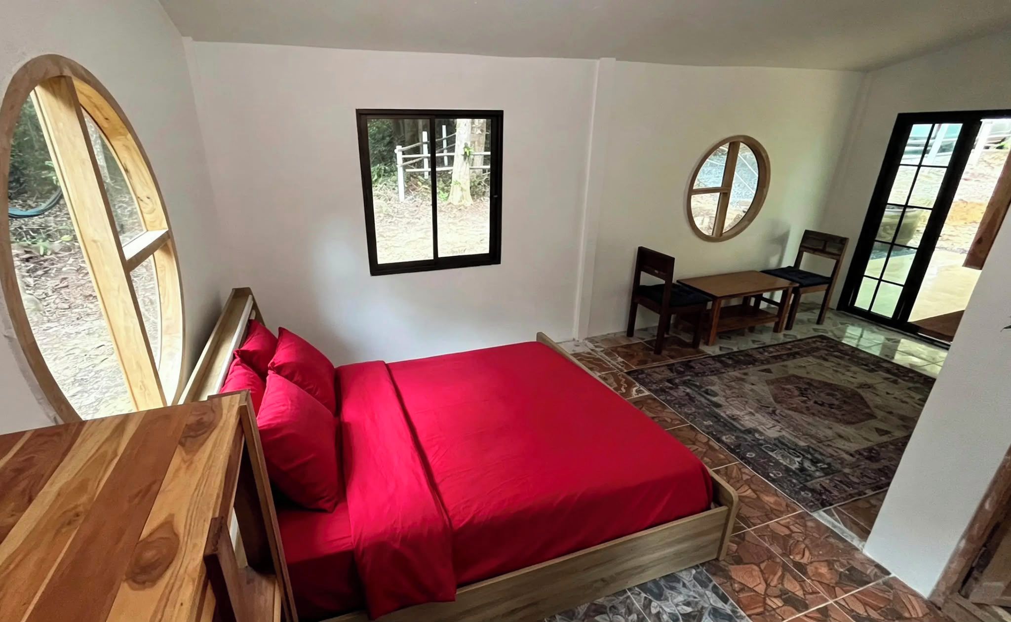 A bedroom with a bed, a desk, a chair, a small table, and a window.