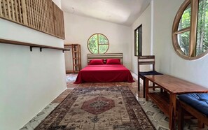 A bedroom with a bed, a wooden chair, a small table, a window with a view of trees, and a patterned rug.