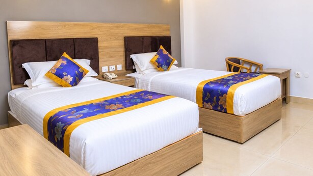 A hotel room with two beds, each with a wooden headboard, white bedding, and a blue and yellow floral bedspread. There is a wooden nightstand with a telephone and a small table with a chair. - Nile View Hotels - Quara  (Gondar)