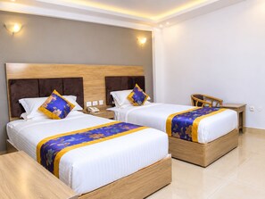 Habitación de hotel con dos camas, cada una con cabecero de madera, ropa de cama blanca y colcha floral azul y amarilla. Hay una mesita de noche con un teléfono y una pequeña mesa con una silla.